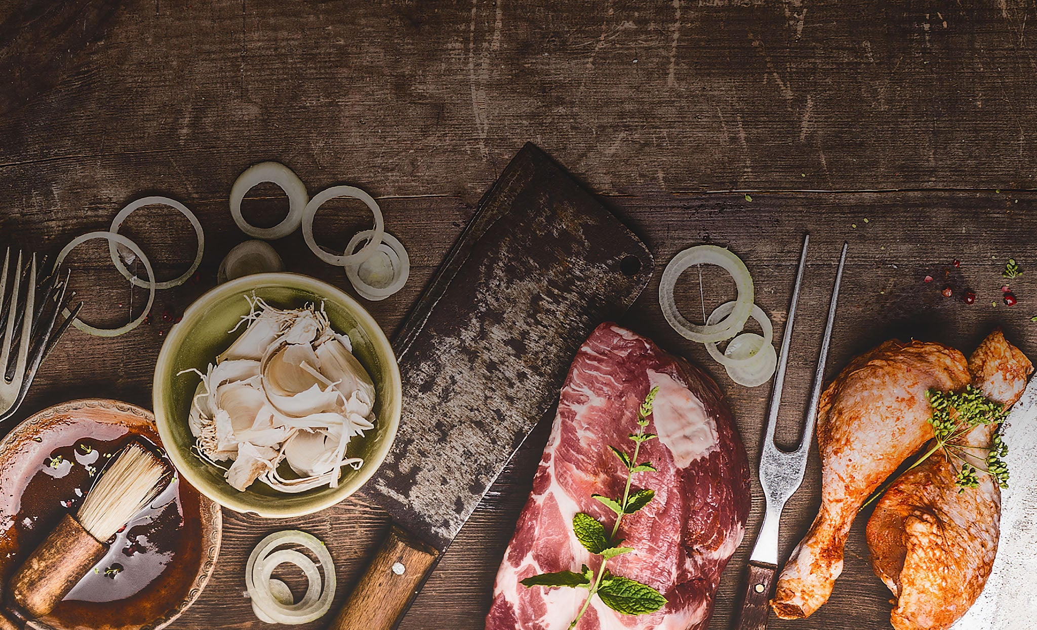 A bowl with a sauce and brush, a bowl full of garlic, a meat cleaver, a cut of steak with fresh herbs, a grilling fork, and chicken drumsticks with fresh herbs, resting on a butcher block and scattered rings of onions.