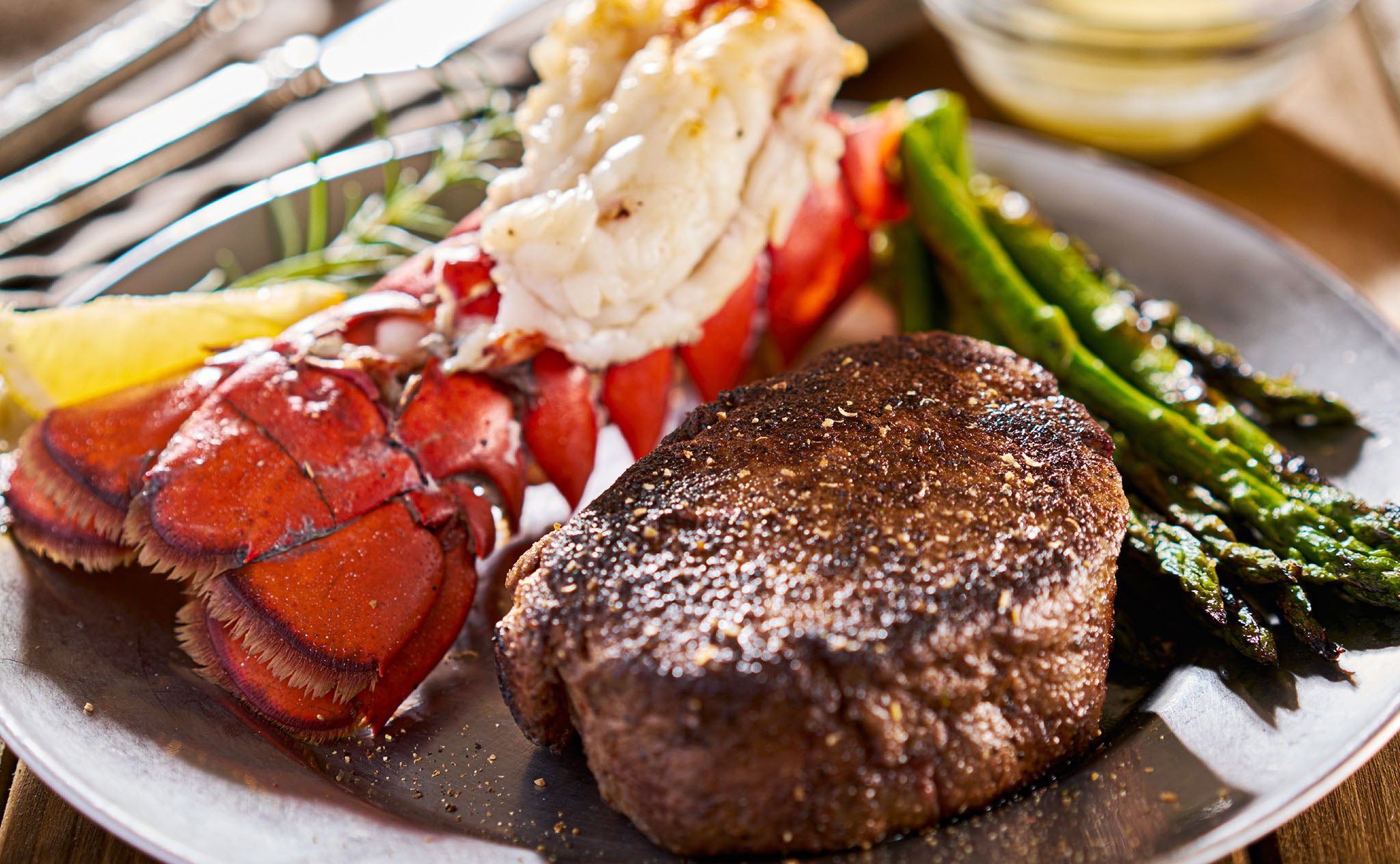 A serving dish with a grilled lobster tail, steak, asparagus, and lemon wedge.